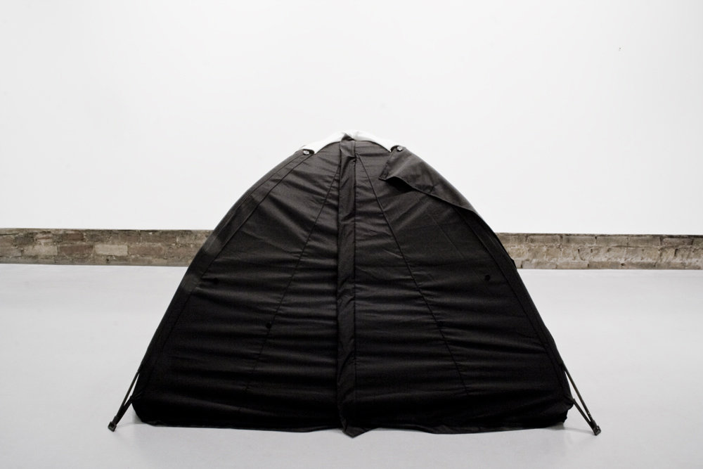 Black textile tent-like object installed indoors, photographed against a white exhibition wall.
