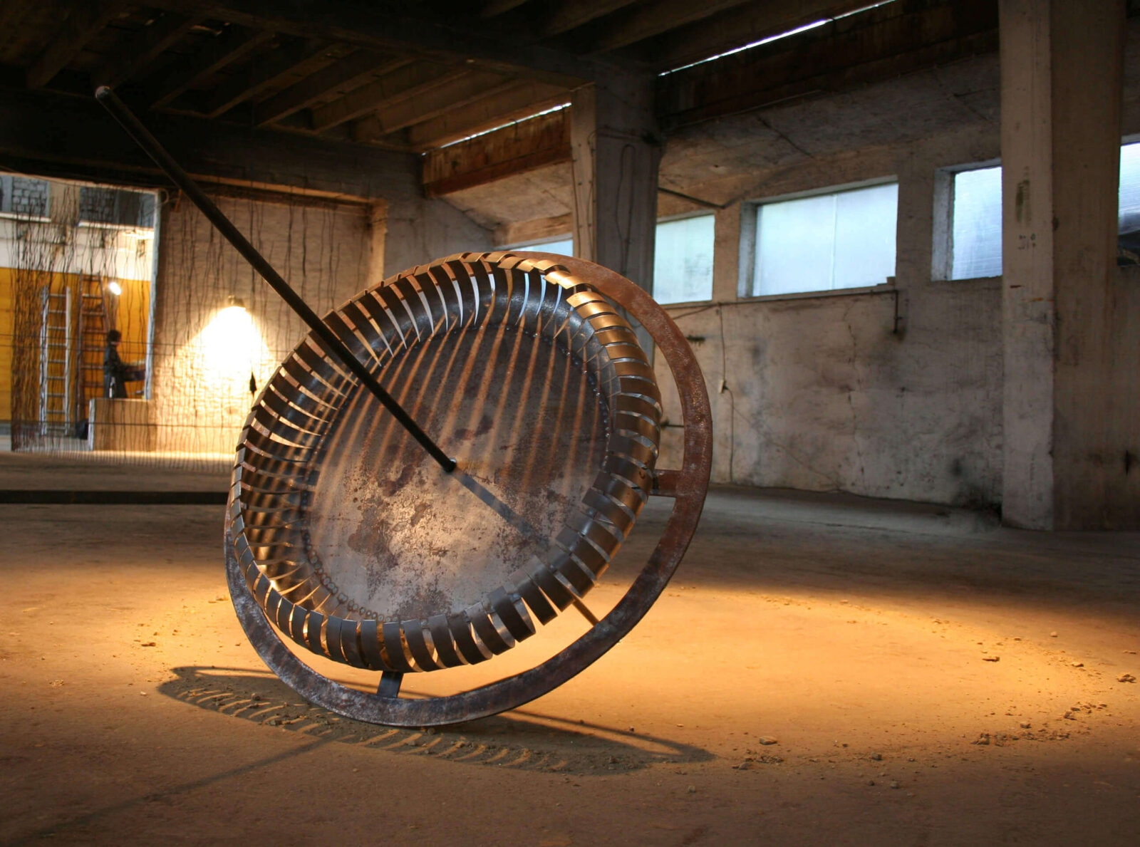 Interactive sculpture consisting of a large circular metal structure positioned on the floor of an industrial interior space.