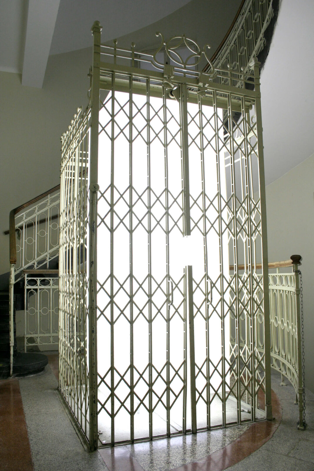 Illuminated cubic light object enclosed by a metal elevator cage in a historic stairwell.