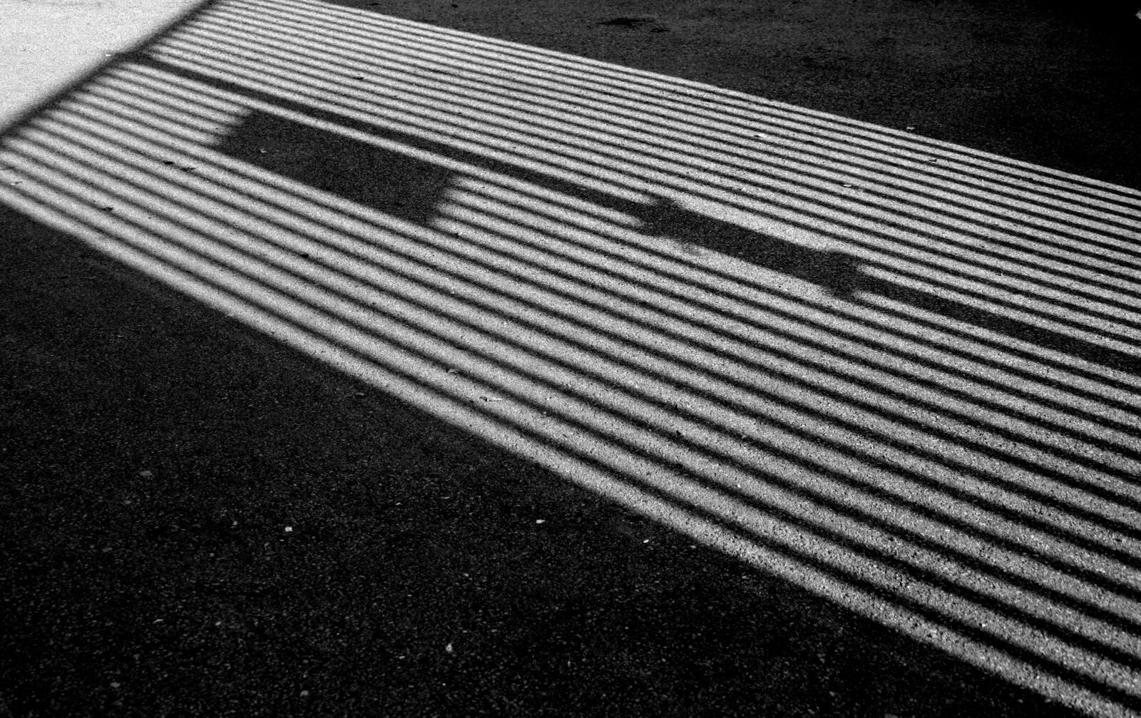 Photograph showing repetitive linear shadows forming a geometric pattern on asphalt.