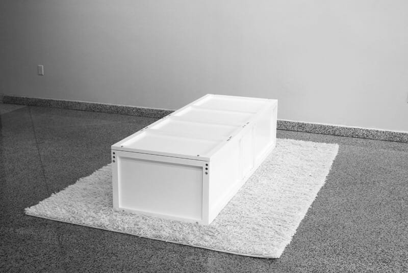 Transformable object, white modular coffin-furniture hybrid structure, closed rectangular form resting on white carpet, exhibited in minimalist gallery space.