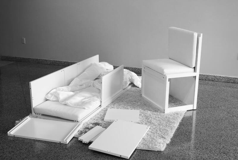 Transformable object shown disassembled into multiple white furniture components, including panels and cushions, arranged on a white rug.
