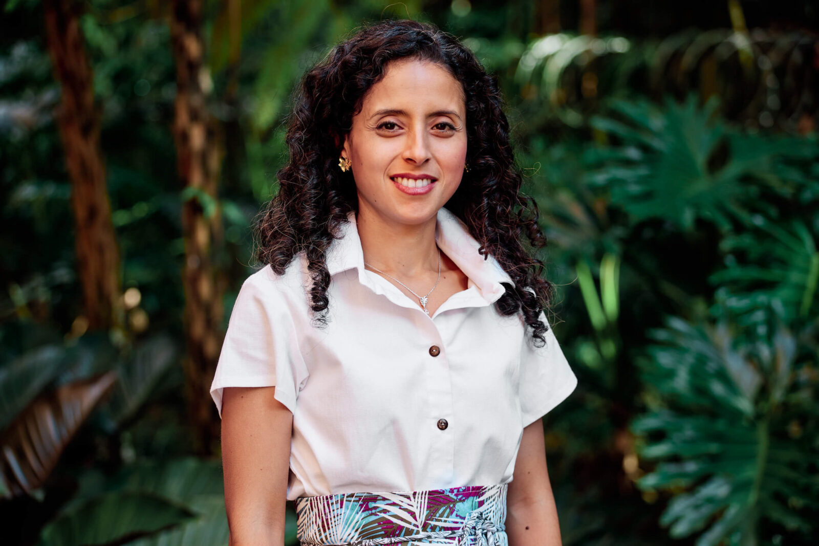 Portrait photograph of a woman standing outdoors in front of green foliage, wearing a light-colored blouse and patterned skirt.