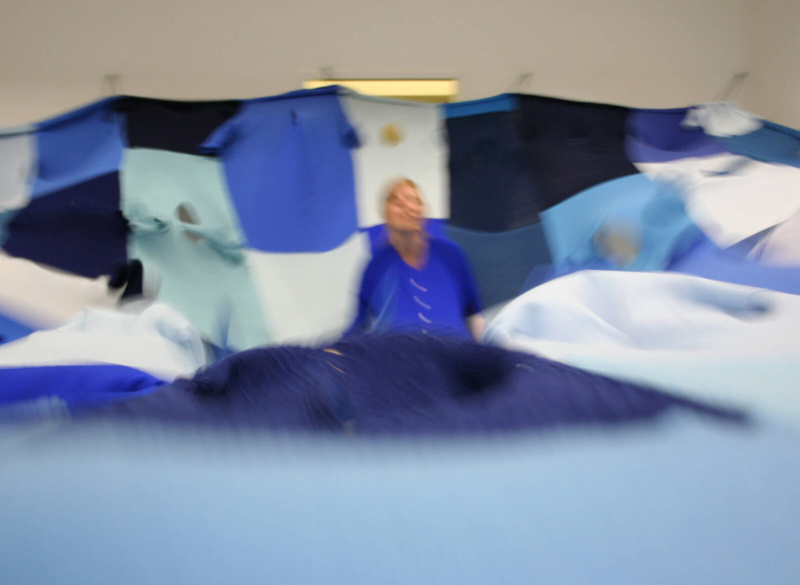 Installation view showing a participant partially obscured by moving blue textile elements suspended overhead in an exhibition space.