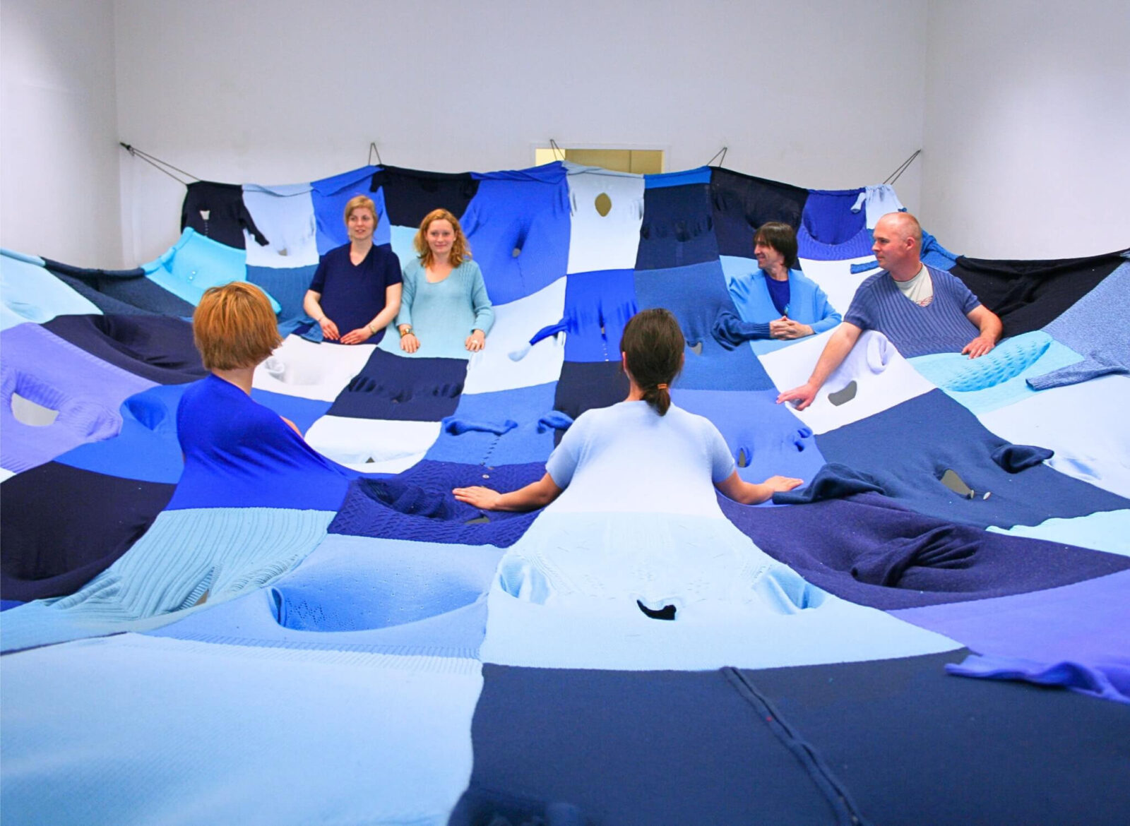 Wide view of an installation where several participants are positioned beneath a large patchwork textile made of blue clothing.