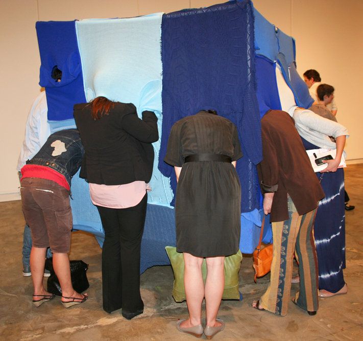 Audience interacting with a blue textile-covered cubic installation, with multiple people bending into openings in the structure.
