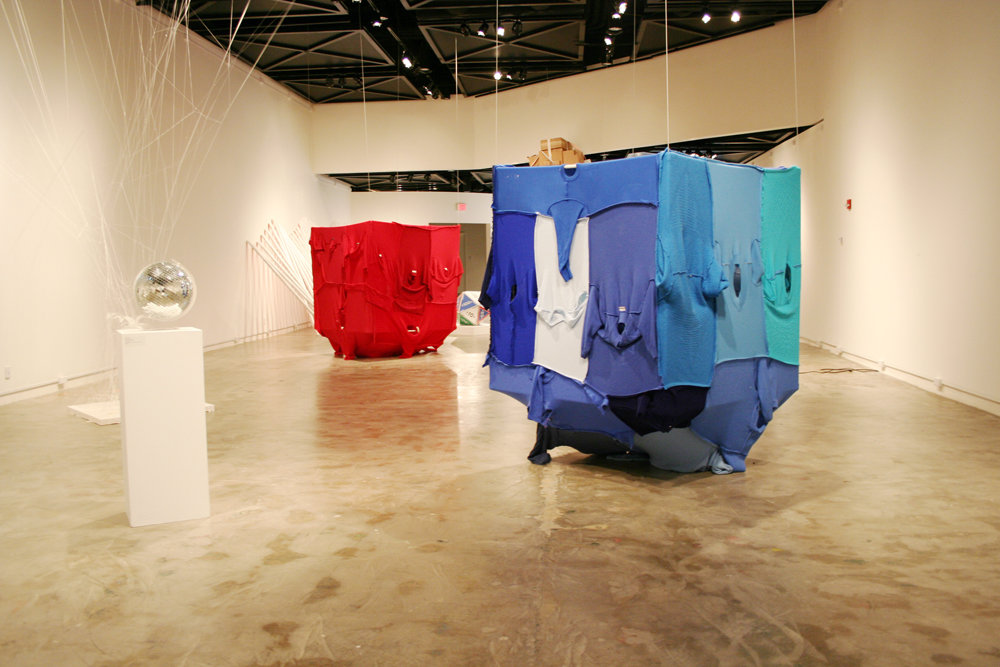 Wide gallery view showing blue and red textile-covered cubic installations suspended and placed within an exhibition space.
