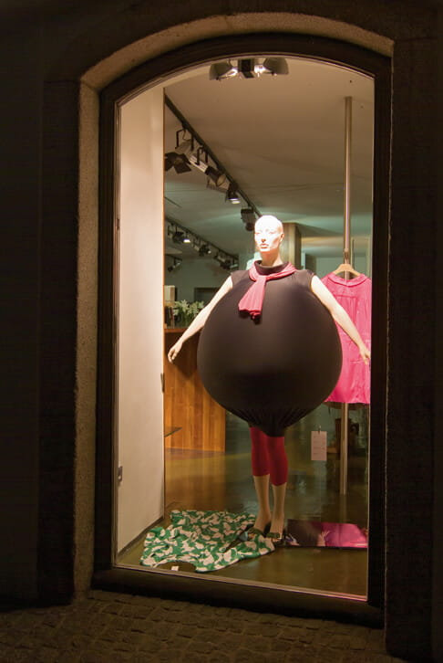 Night view of a storefront installation featuring a mannequin wearing a large black expandable garment positioned in a shop window.
