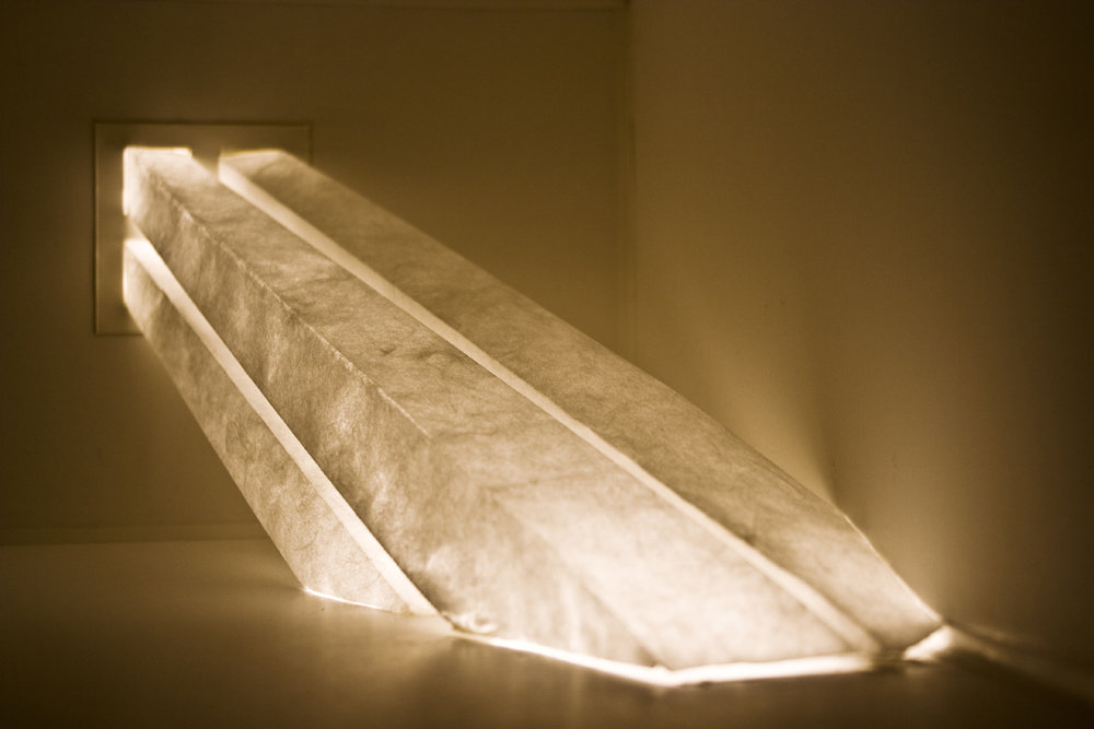 Close-up view of a translucent elongated sculpture emitting warm light from a window to the floor within a darkened room.