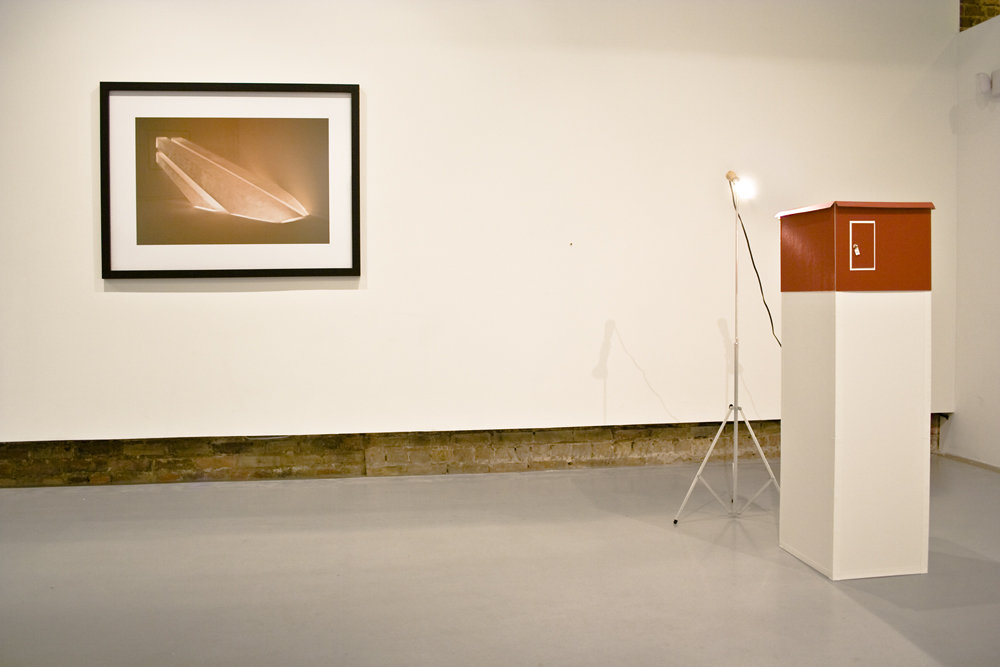 Exhibition view showing a framed photograph on the wall, a lit sculptural object on a pedestal, and a spotlight in a gallery space.