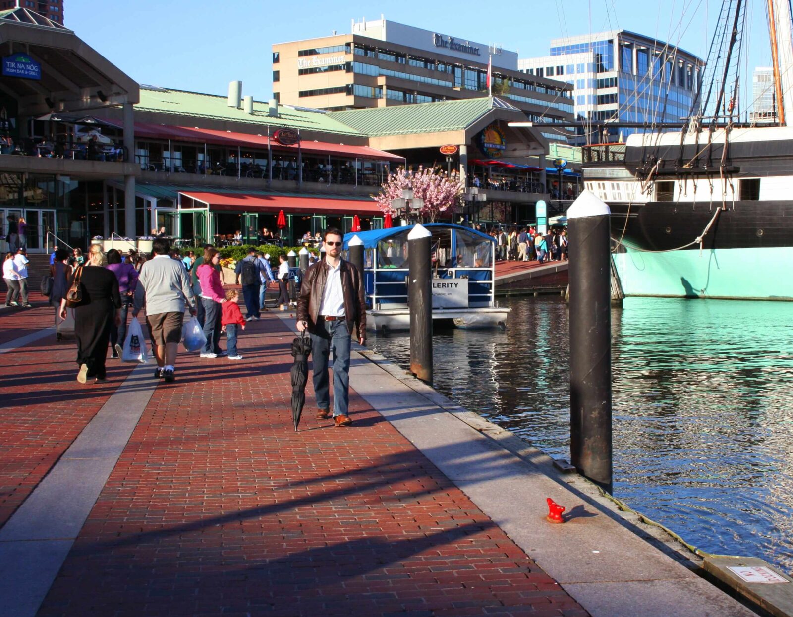 Man walking along a waterfront promenade of Baltimore USA holding a closed umbrella, with people, buildings, and boats visible in the background.