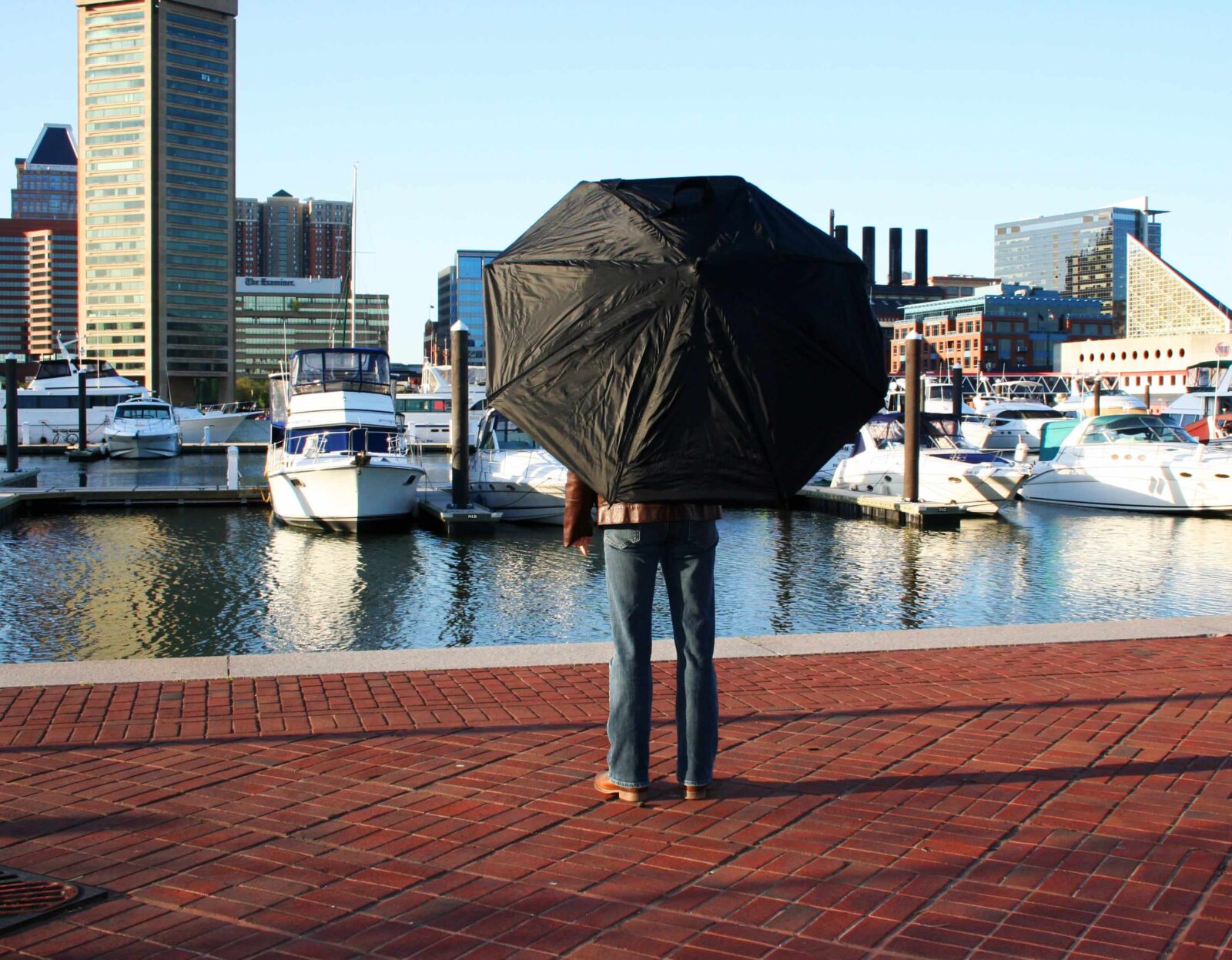Person standing on a brick waterfront promenade of Baltimore USA holding an open black umbrella, facing a marina with boats and city buildings.