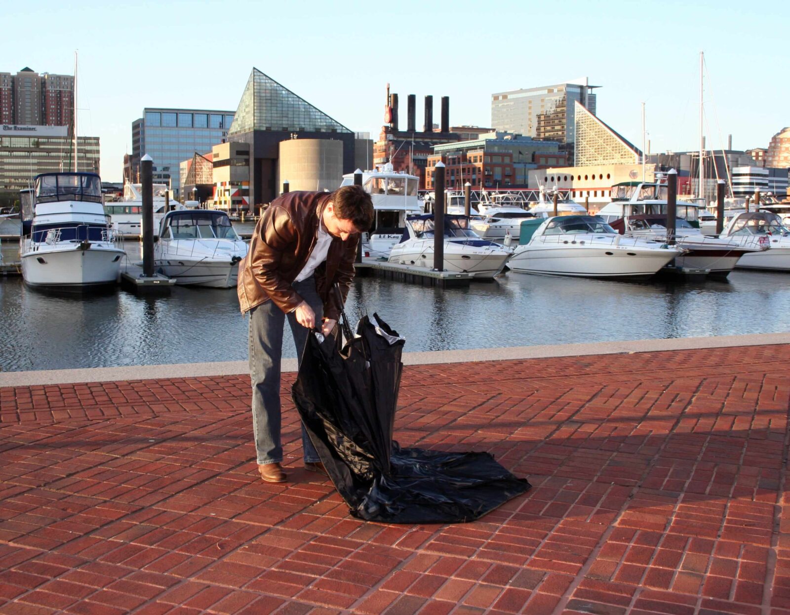 Person bending down on a brick promenade of Baltimore USA while folding or unfolding a black umbrella, with boats and urban waterfront architecture in the background.