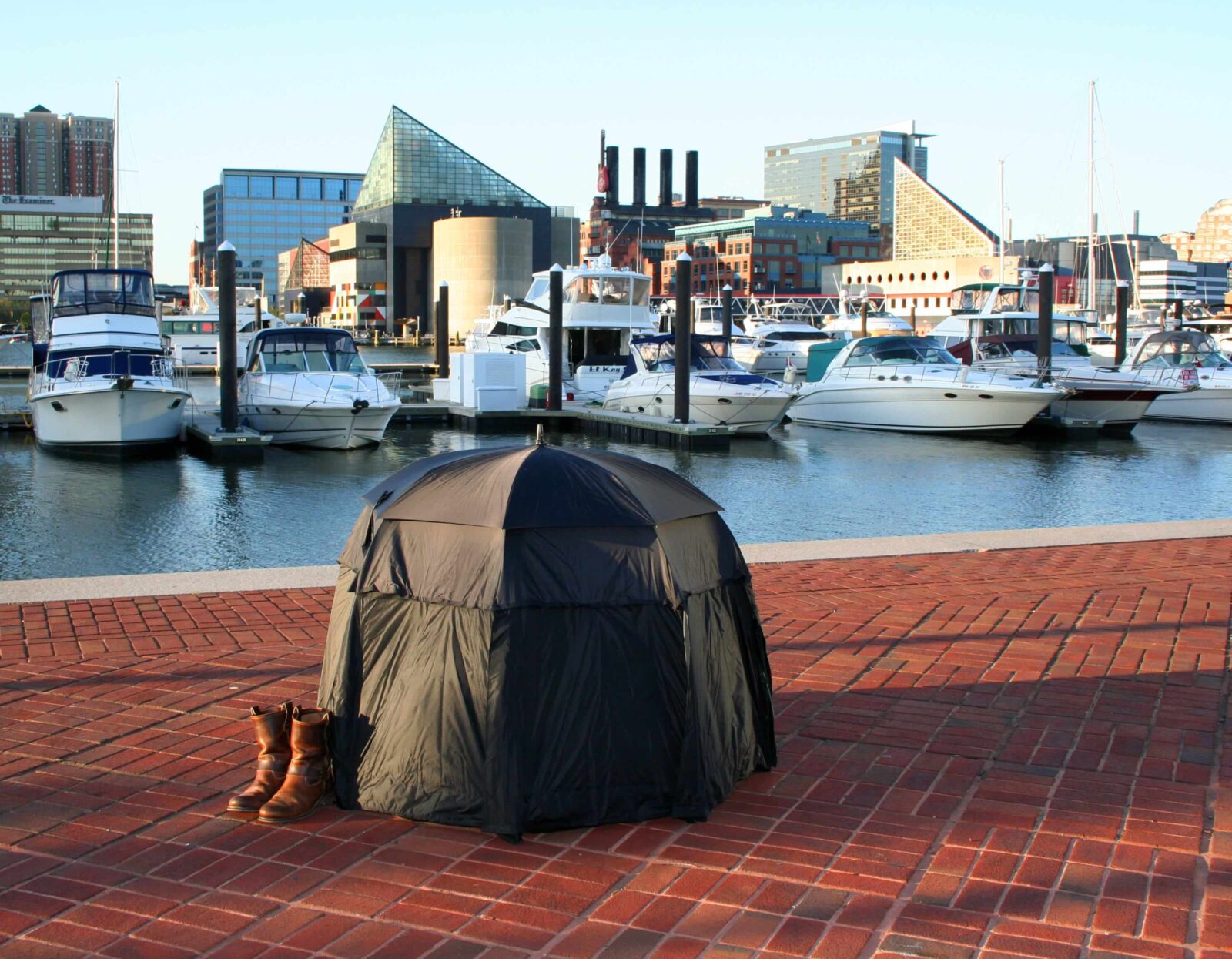 Black umbrella transformed into a dome-like textile structure placed on a brick promenade of Baltimore USA beside a waterfront, with shoes positioned next to it.