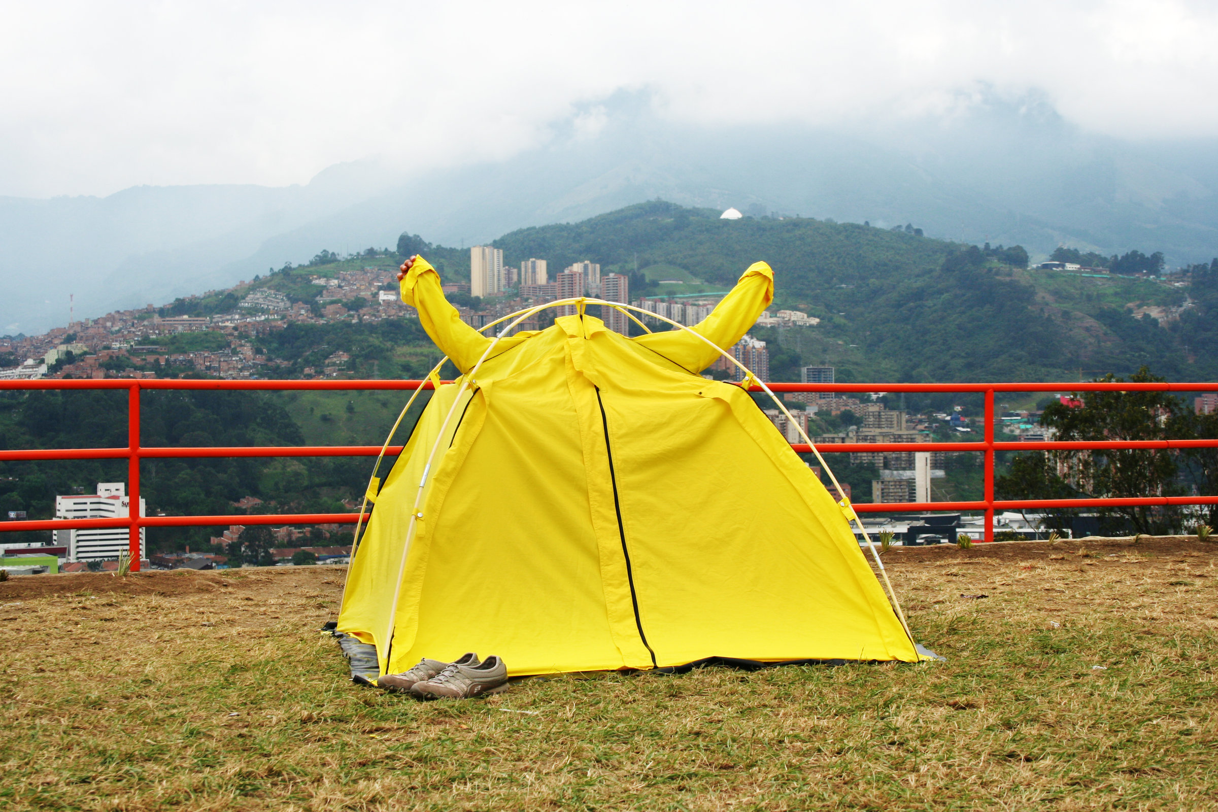 Performance textile object, yellow tent-shelter fully erected on hillside platform in Medellín, arms raised in the sleeves while tent closed with shoes visible at entrance, photographed against mountainous urban landscape.