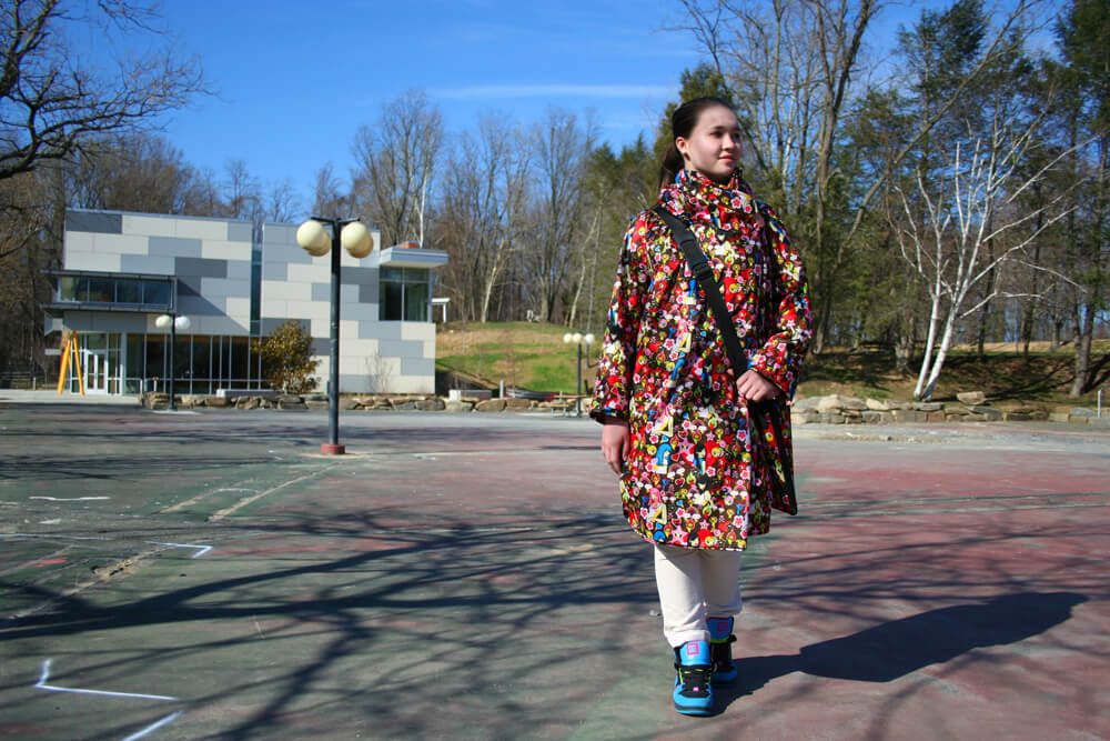 Performance documentation showing a person wearing a multicolored patterned textile garment standing in an outdoor public space with modern buildings in the background.