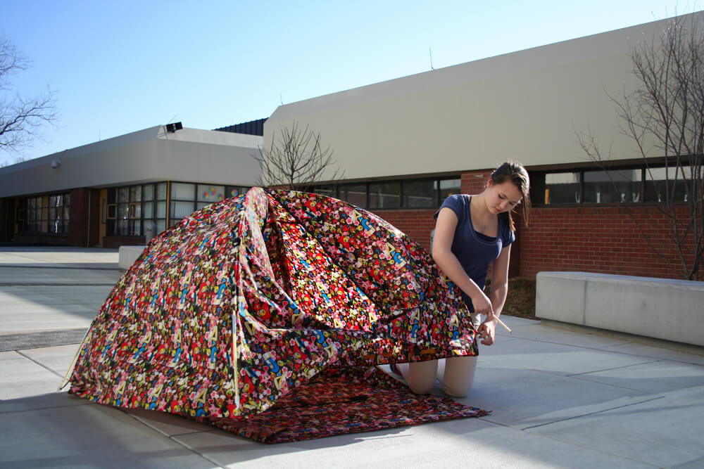 Textile object installation showing a dome-shaped structure made of multicolored patterned fabric being assembled by a person in an outdoor courtyard setting.
