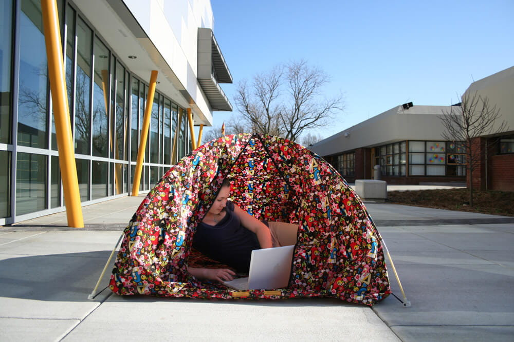 Textile object installation showing a person inside a dome-shaped structure made of multicolored patterned fabric, using a laptop in an outdoor architectural environment.