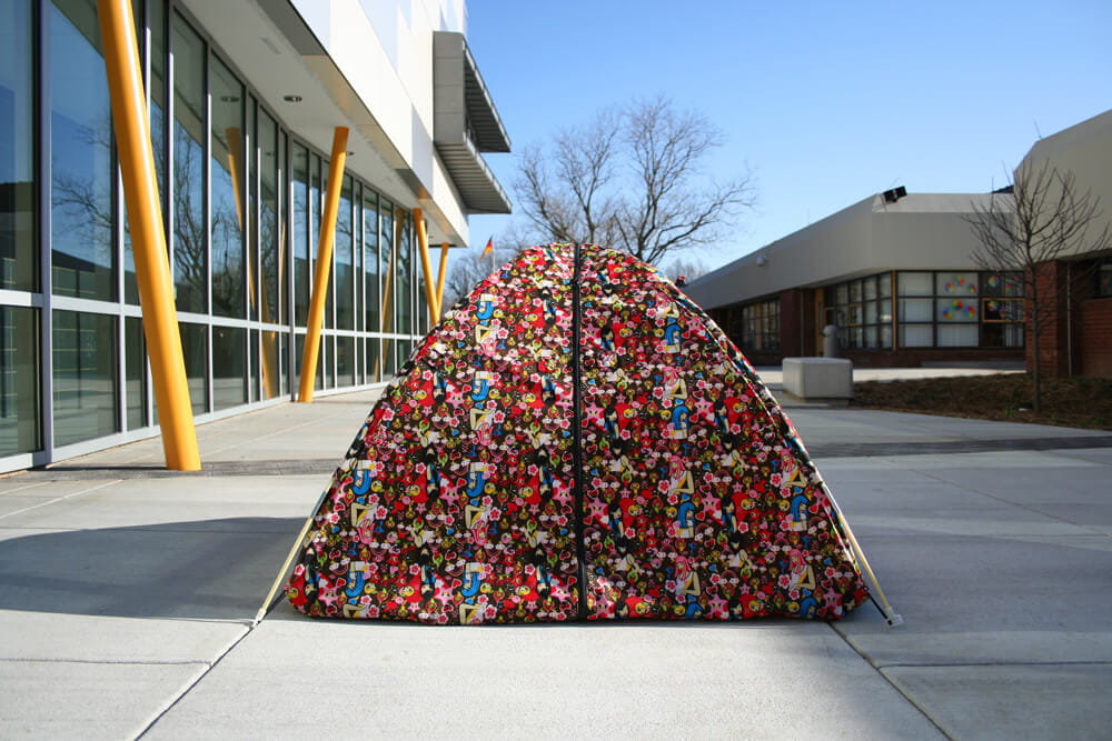 Textile sculpture, patterned tent made of colorful fabric, standing assembled, photographed on an outdoor concrete plaza in front of a modern building.