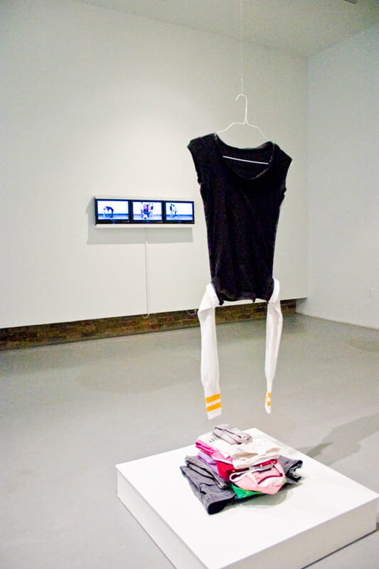 Installation, black shirt and white socks hanging from a wire henger, suspended in mid-air above a stack of clothes, photographed in a gallery space.