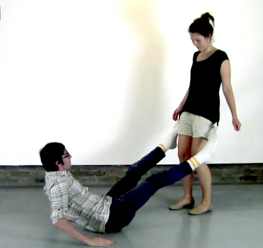 Performance textile collage, male performer sitting on floor with open legs and standing female performer physically connected by zippered fabric attachments linking feet via socks and hips via t-shirt, performing interdependent movement in white gallery space with industrial floor.