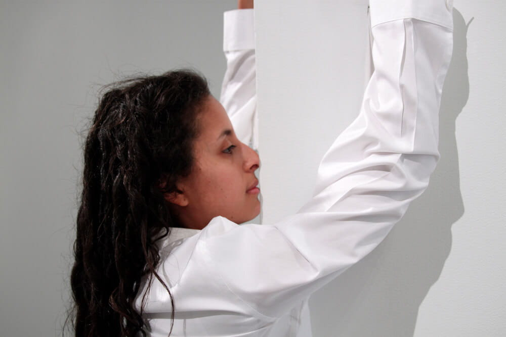 Performance art, woman in a white shirt, facing against a white corner with arms raised, photographed in a close-up profile view.