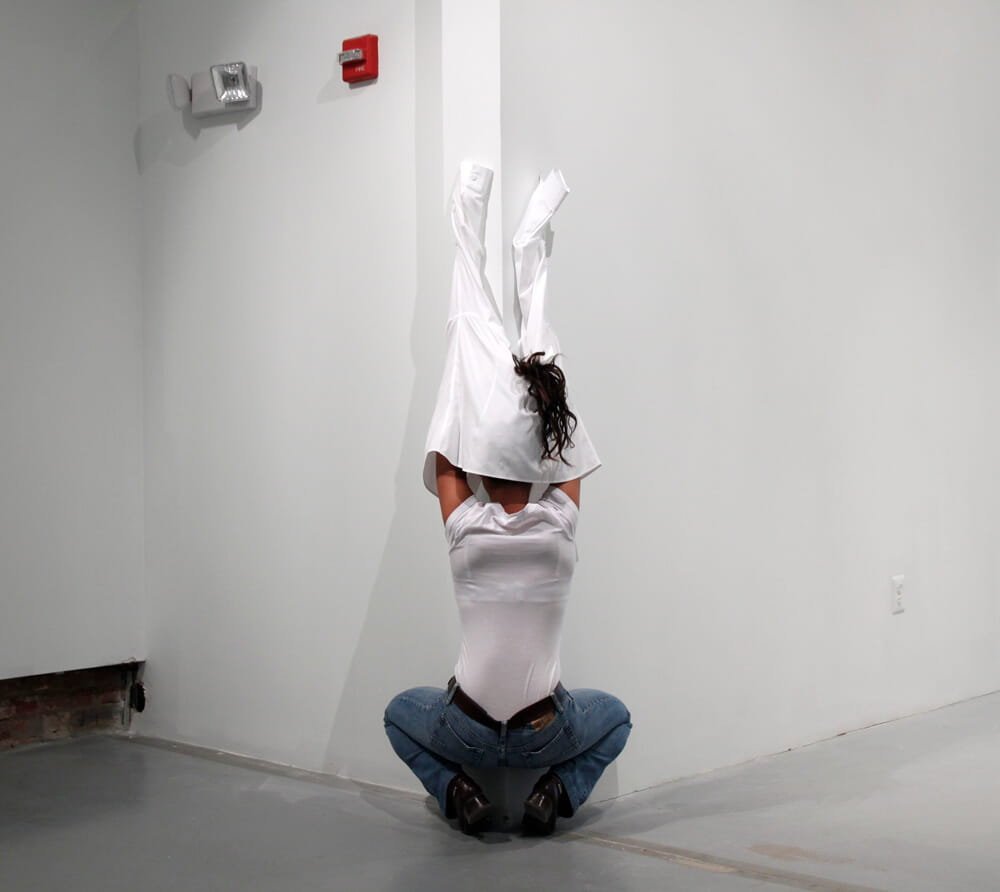 Performance art, woman in a white shirt and jeans, crouching facing at a wall corner with her shirt pulled over her head and attached to the wall, photographed from behind.