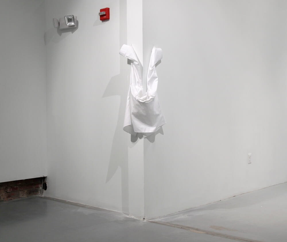 Installation, white shirt attached to a wall corner by its sleeves, hanging empty in a draped position, photographed in a white gallery space.