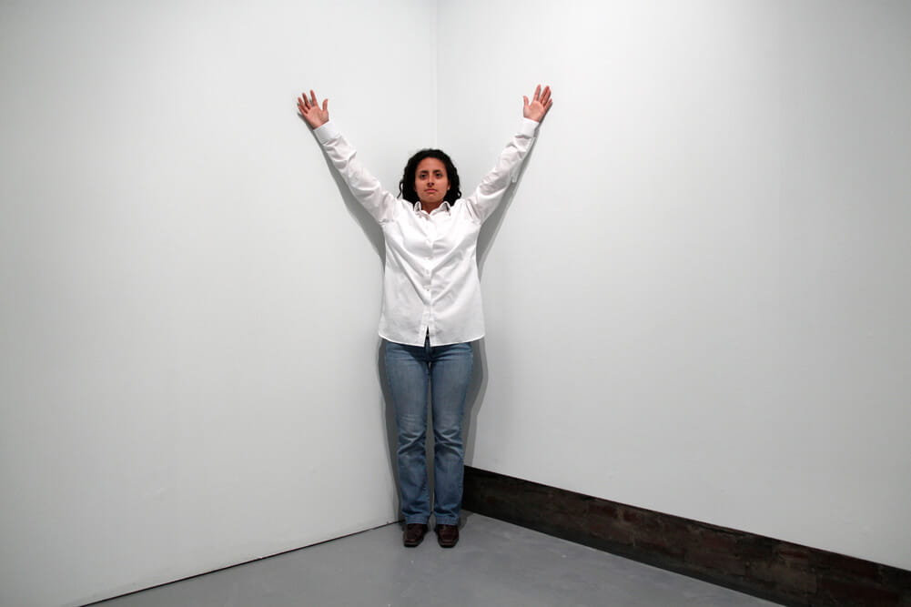 Performance art, woman in a white shirt and jeans, standing in a wall corner with arms raised in a V-shape, photographed frontally in a gallery.