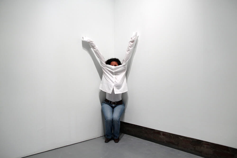Performance art, woman in a white shirt and jeans, crouching in a corner with her shirt pulled up over her face and attached to the walls, photographed in a white room.