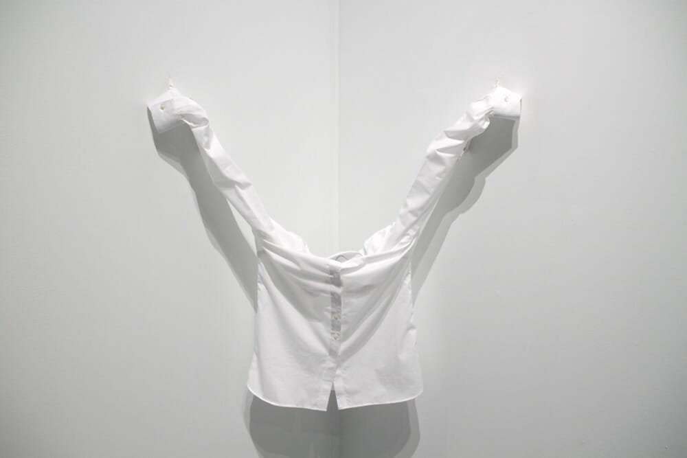 Installation, white button-down shirt, pinned to a wall corner by its extended sleeves to form a V-shape, photographed as a static object in a gallery.